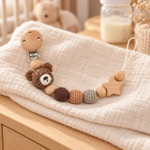 Schnullerkette mit gehäkeltem Bär-Motiv, Holzperlen, Holzstern und rundem Holzclip auf hellem Musselintuch.
